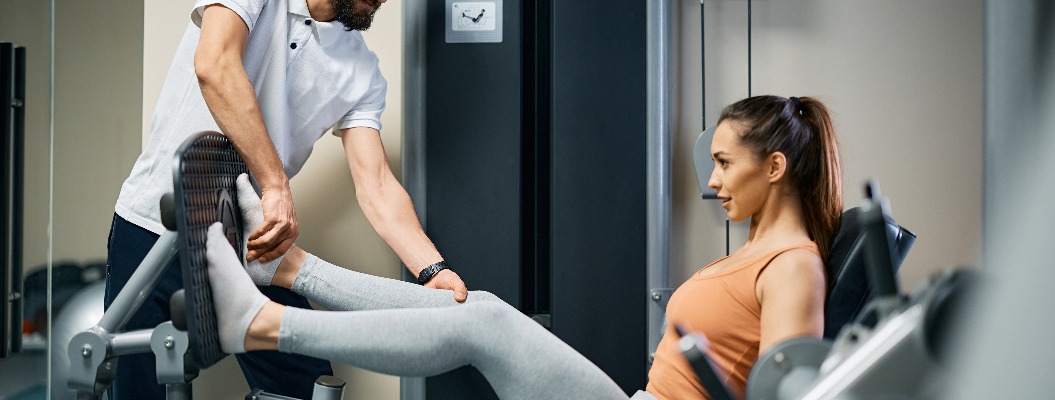 Junge Sportlerin trainiert mit Hilfe eines Physiotherapeuten im Fitnessstudio an der Beinpresse.