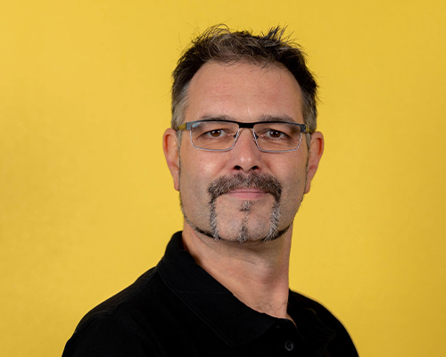 Ein Mann mit Brille und Schnurrbart laechelt freundlich vor gelbem Hintergrund