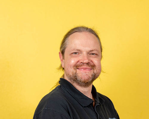 Ein lächelnder Mann mit langen Haaren und Bart vor gelbem Hintergrund in dunklem Shirt