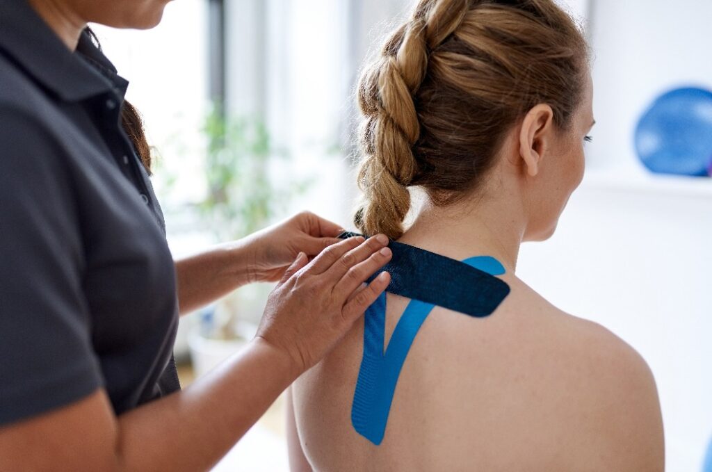 Eine chinesische Massagetherapeutin bringt Kinesio-Tape an den Schultern und am Nacken einer attraktiven blonden Kundin in einer hellen Arztpraxis an.